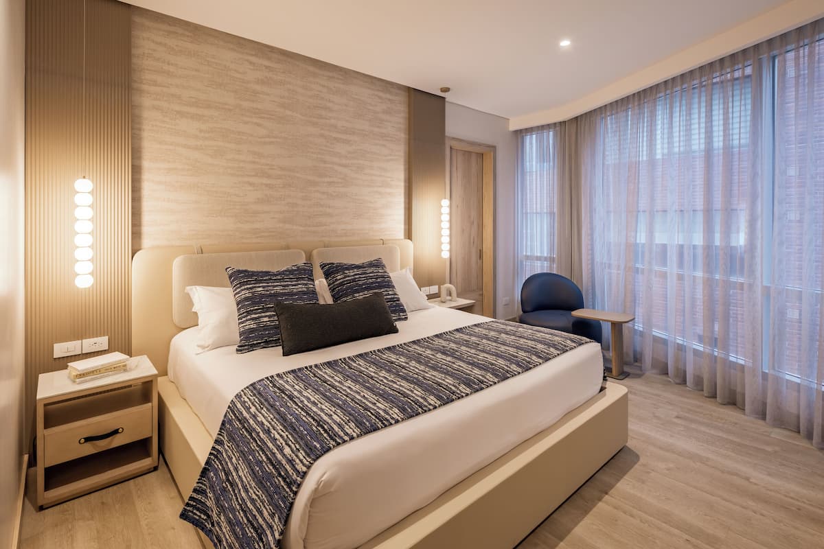 Un dormitorio de hotel moderno con una cama grande, mesitas de noche, una silla y vistas a la ciudad a través de amplios ventanales.