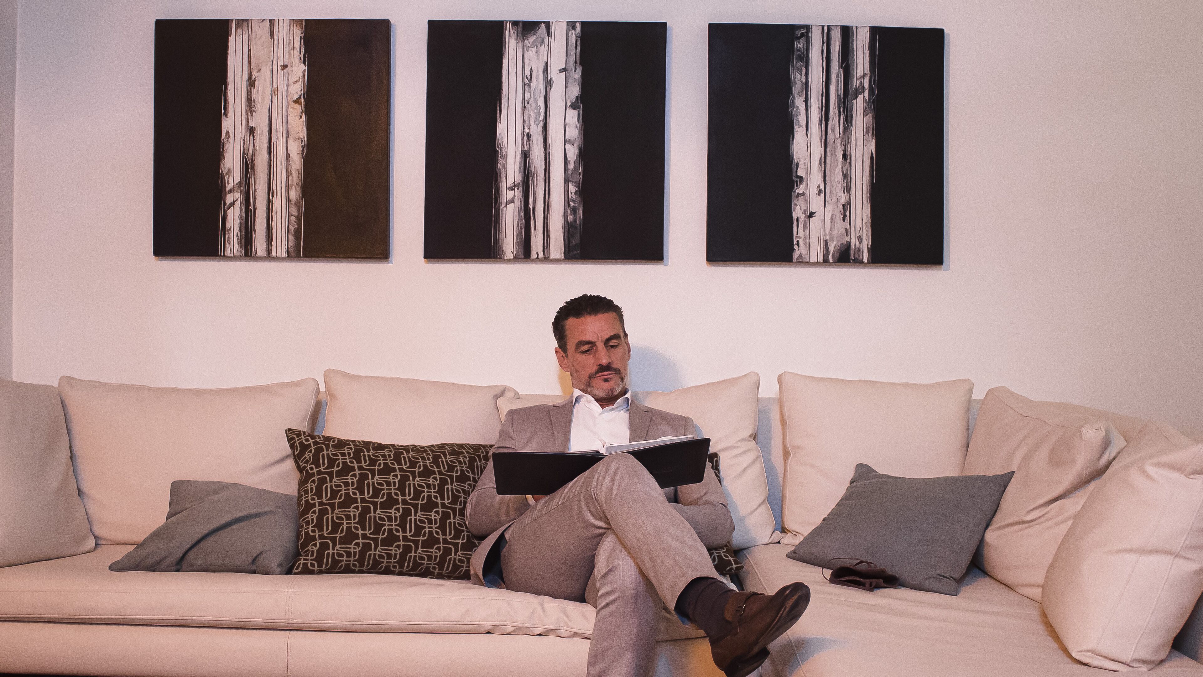 A man sitting on a beige sofa with a book, surrounded by patterned pillows, in a room with abstract paintings on the wall.
