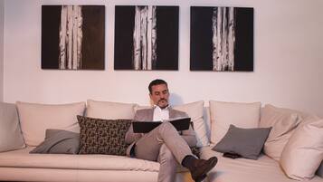 A man sitting on a beige sofa with a book, surrounded by patterned pillows, in a room with abstract paintings on the wall.