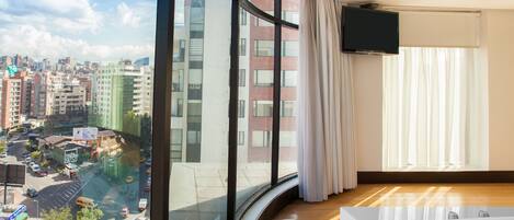 A hotel room with a large window offering a city view, a flat-screen TV mounted on the wall, and a wooden floor.