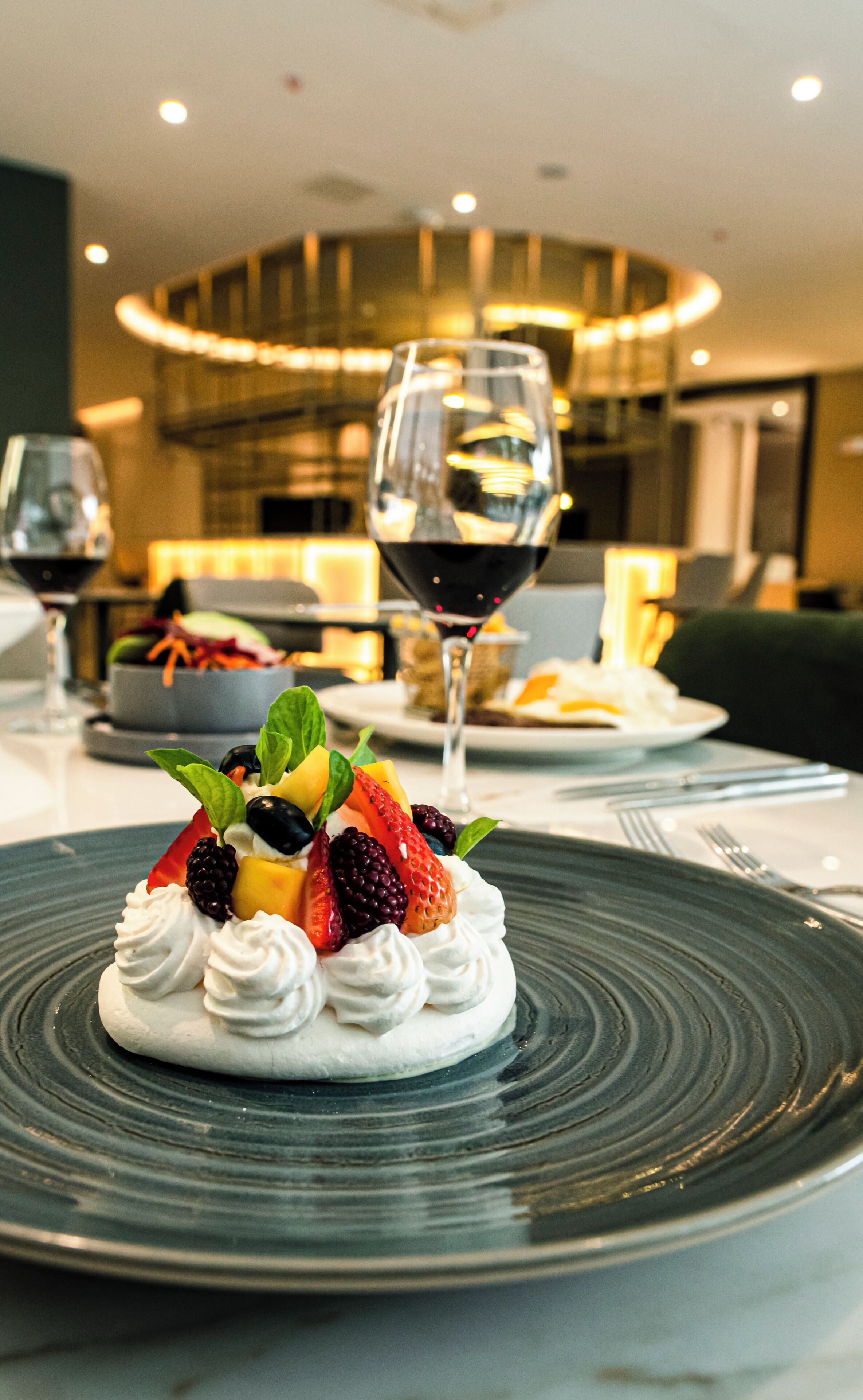 A dessert on a plate with whipped cream and berries, a glass of red wine, and a bowl of salad in the background.