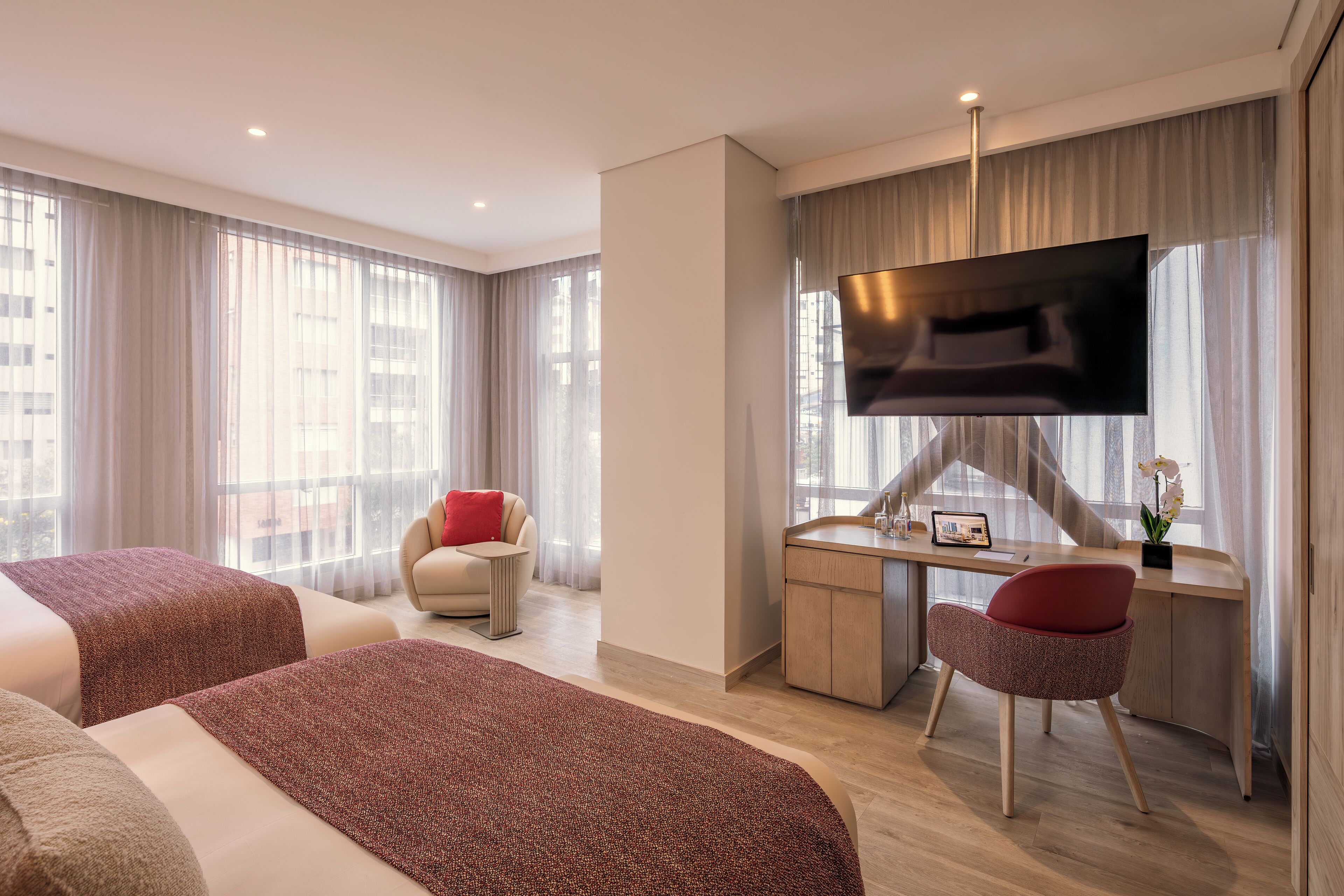 A hotel room with two beds, a desk with a TV, a chair, and a view of buildings through large windows.