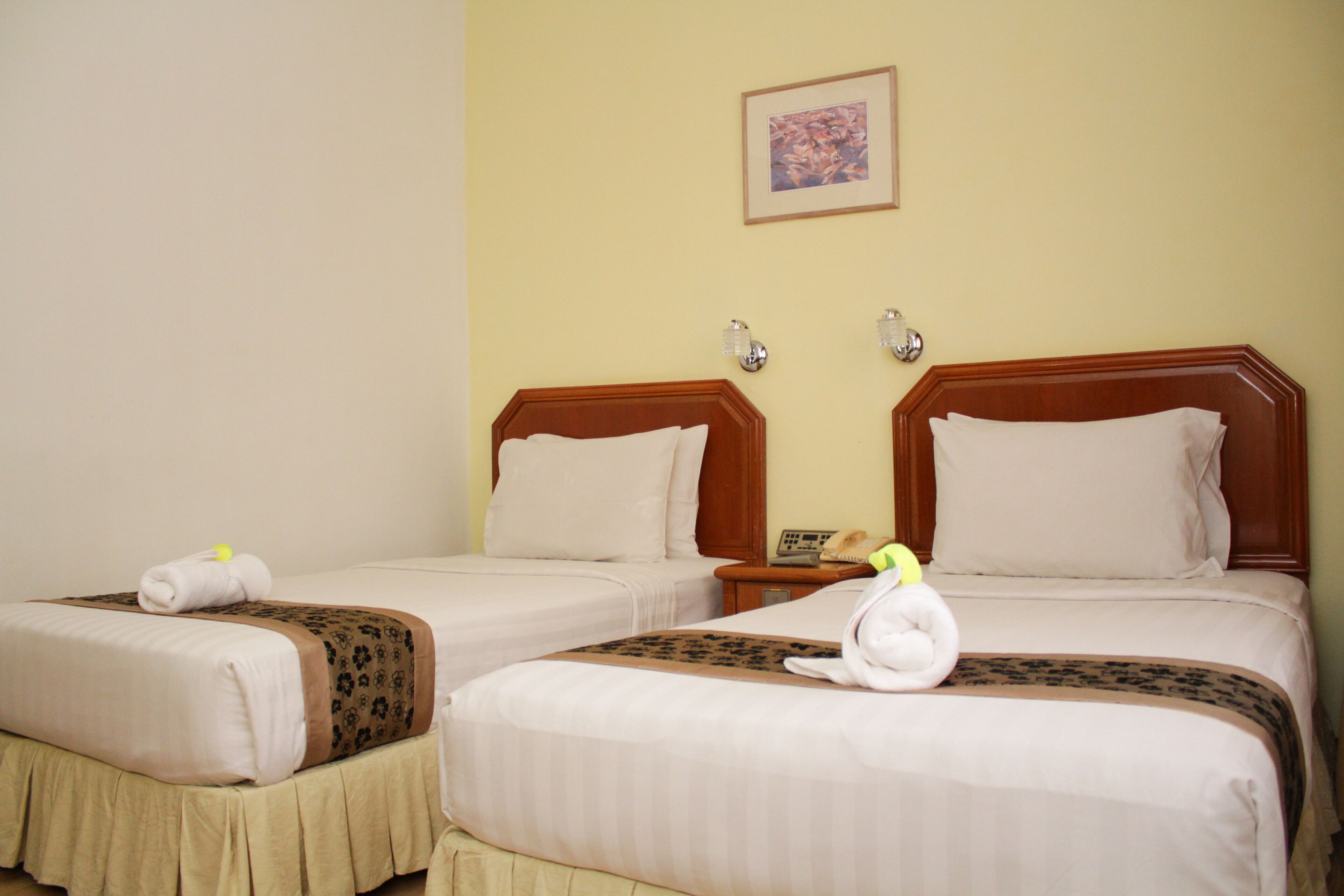 A hotel room with two beds, each with a wooden headboard, white linens, and a brown decorative border. There are two wall-mounted lamps and a framed picture above the beds.