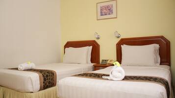 A hotel room with two beds, each with a wooden headboard, white linens, and a brown decorative border. There are two wall-mounted lamps and a framed picture above the beds.