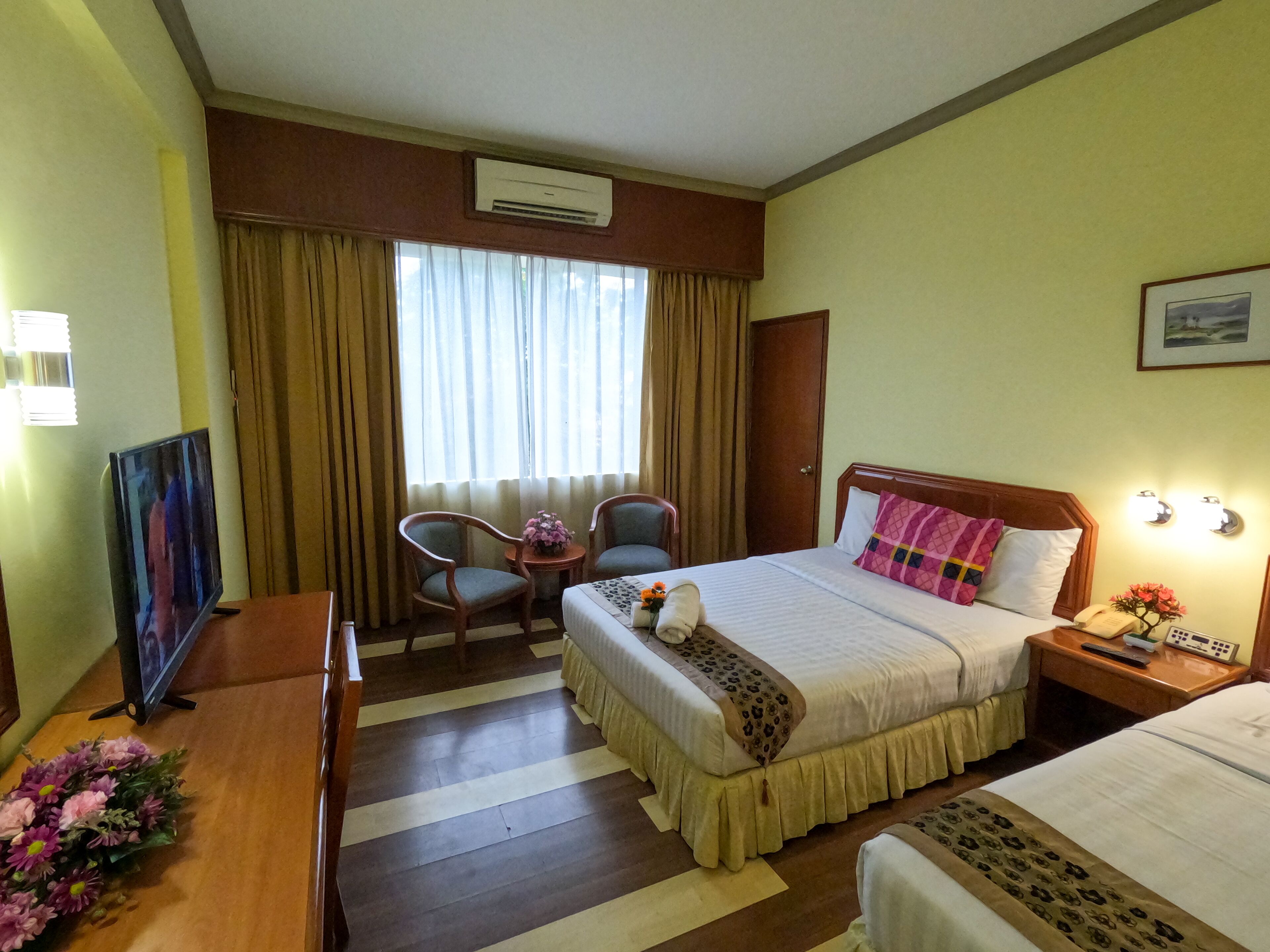 A hotel room with a bed, a desk with a TV, two chairs, a small table with flowers, and a window with curtains.