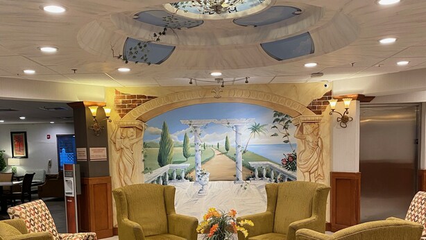 Eine Lobby mit Wandgemälde einer Landschaft, grünen Polsterstühlen, einem Marmor-Couchtisch und einem gemusterten Teppich.