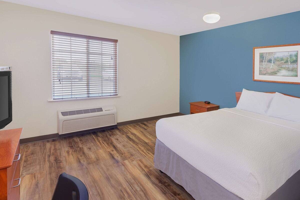Habitación de hotel con cama, escritorio, silla, ventana con persianas, calefactor y un cuadro en la pared.