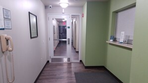A hallway with a wooden floor, a telephone on the left, and a window with blinds on the right. - WoodSpring Suites Ankeny Des Moines (Ankeny)