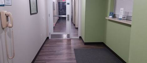 A hallway with a wooden floor, a telephone on the left, and a window with blinds on the right.