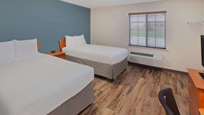 A hotel room with two beds, a desk, a chair, a window with blinds, and a wall-mounted air conditioning unit. - WoodSpring Suites Ankeny Des Moines (Ankeny)