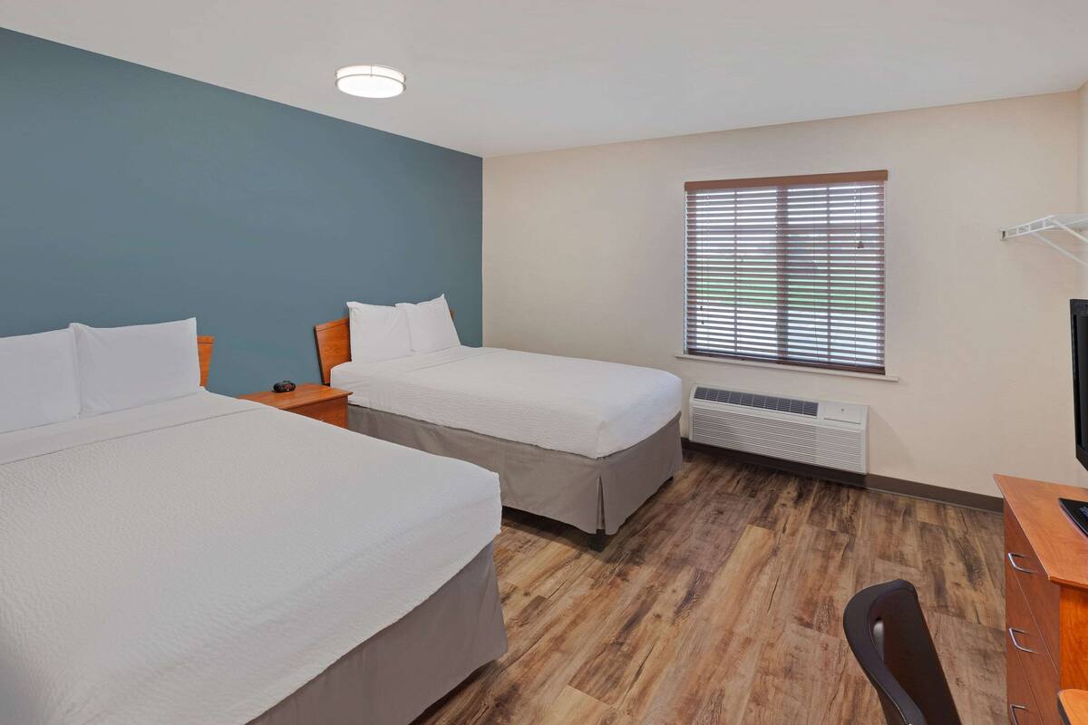 Habitación de hotel con dos camas, un escritorio, una silla, un ventanal con persianas y un aire acondicionado de pared.