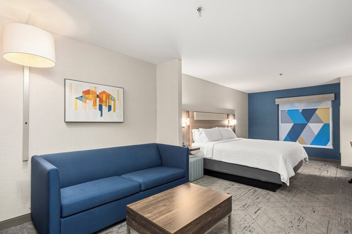 A hotel room with a blue sofa, a wooden coffee table, a bed with white bedding, a nightstand, a lamp, and a geometric wall art piece.