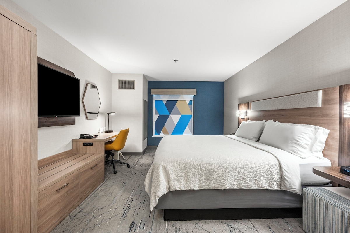 A modern hotel room with a large bed, a desk with a chair, a TV, and a decorative wall panel.