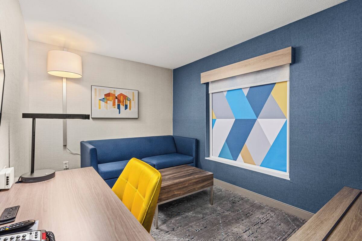 A modern room with a blue sofa, a yellow chair, a wooden coffee table, a lamp, and a geometric wall art piece.