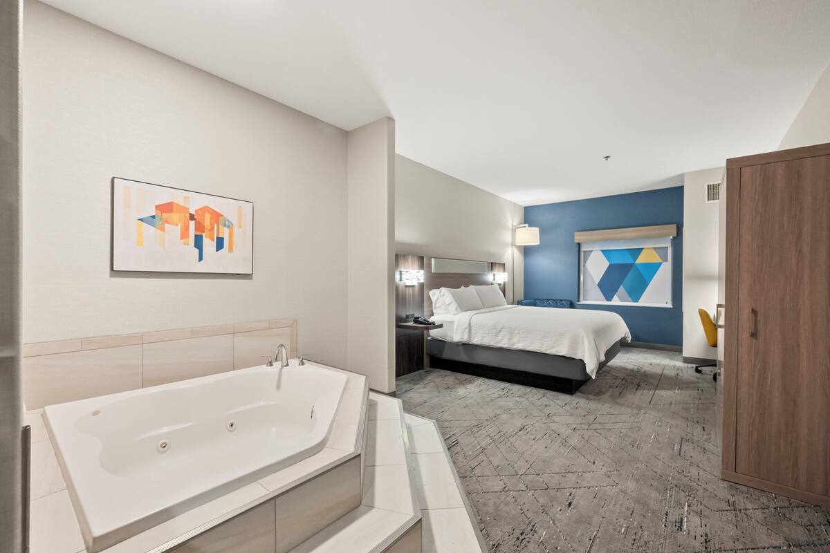 A modern hotel room with a large bathtub, a neatly made bed, a desk with a chair, and a wardrobe.