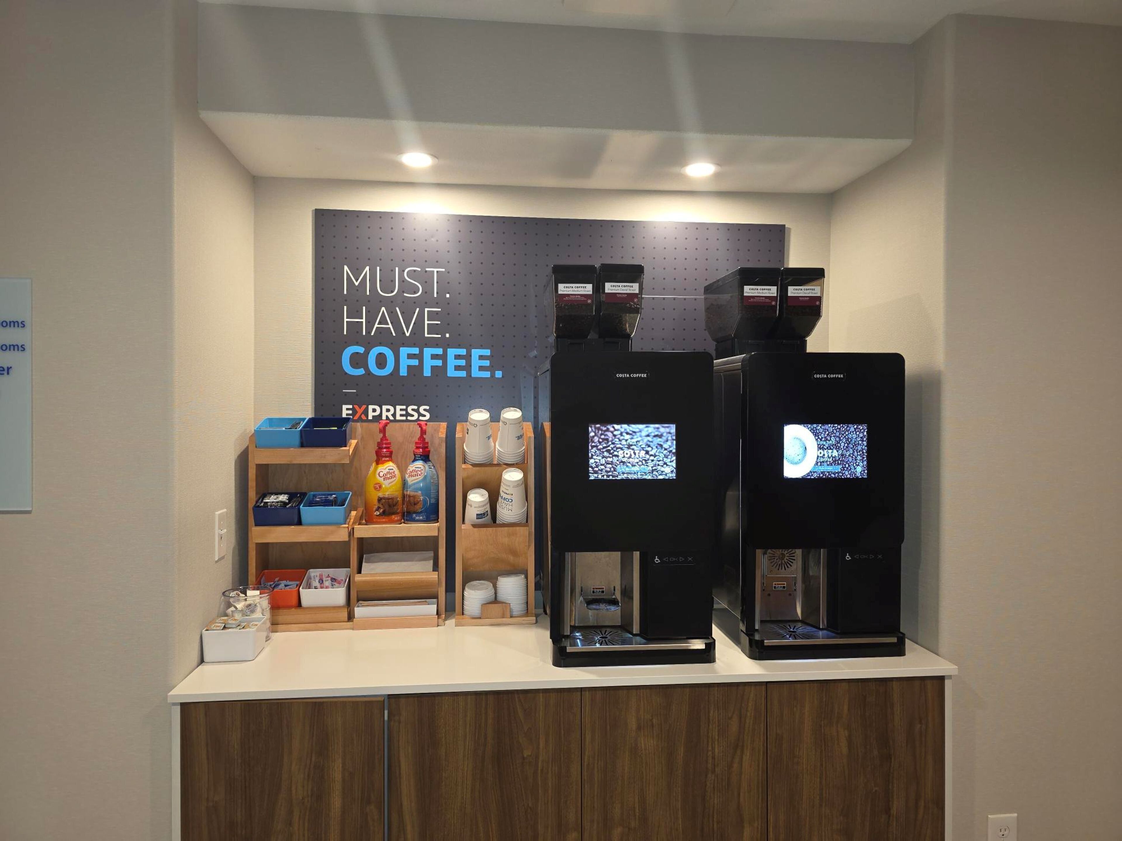 Eine Kaffeestation mit zwei Espressomaschinen, beschriftet mit 'Costa Coffee', und einem Schild mit der Aufschrift 'KAFFEE MUSS SEIN'.