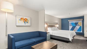 Habitación de hotel con sofá azul, una mesa de centro de madera, cama con ropa blanca, mesita de noche, lámpara y una obra de arte mural geométrica.