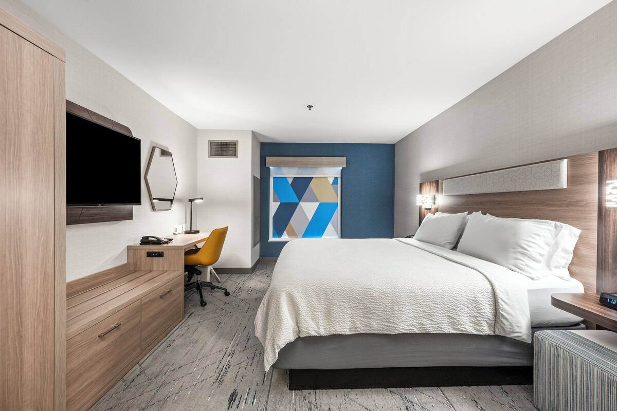 A modern hotel room with a large bed, a desk with a chair, a TV, and a decorative wall panel.