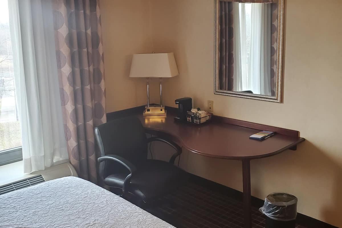 A hotel room with a bed, a desk with a lamp, a chair, a trash can, and a window with curtains.