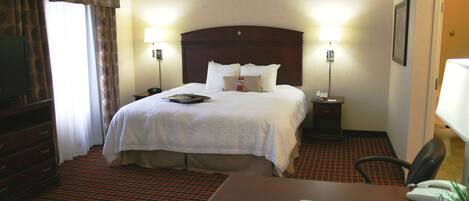 A hotel room with a large bed, two bedside tables, a desk with a chair, a television, and a red and black patterned carpet.