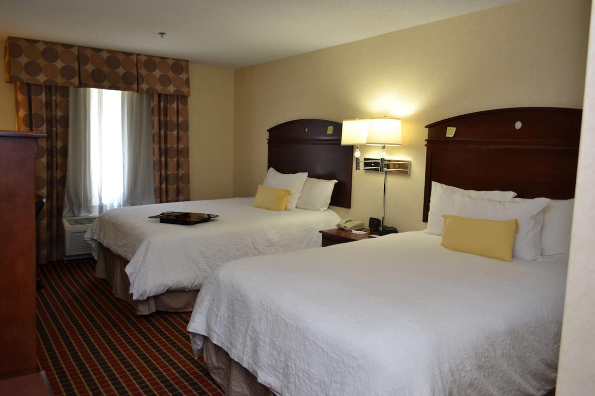 A hotel room with two beds, a large window with curtains, a bedside table with a lamp, and a carpeted floor.