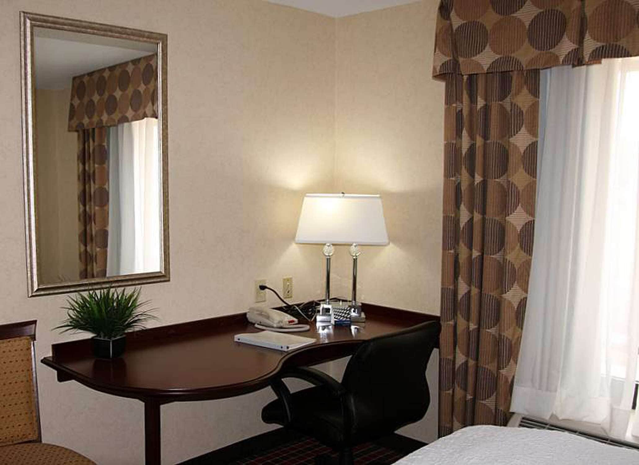 A hotel room with a desk, a chair, a bed, a lamp, a telephone, and a mirror.