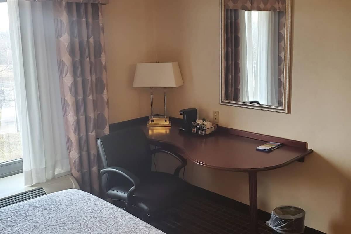 A hotel room with a bed, a desk, a chair, a lamp, a trash can, and a window with curtains.