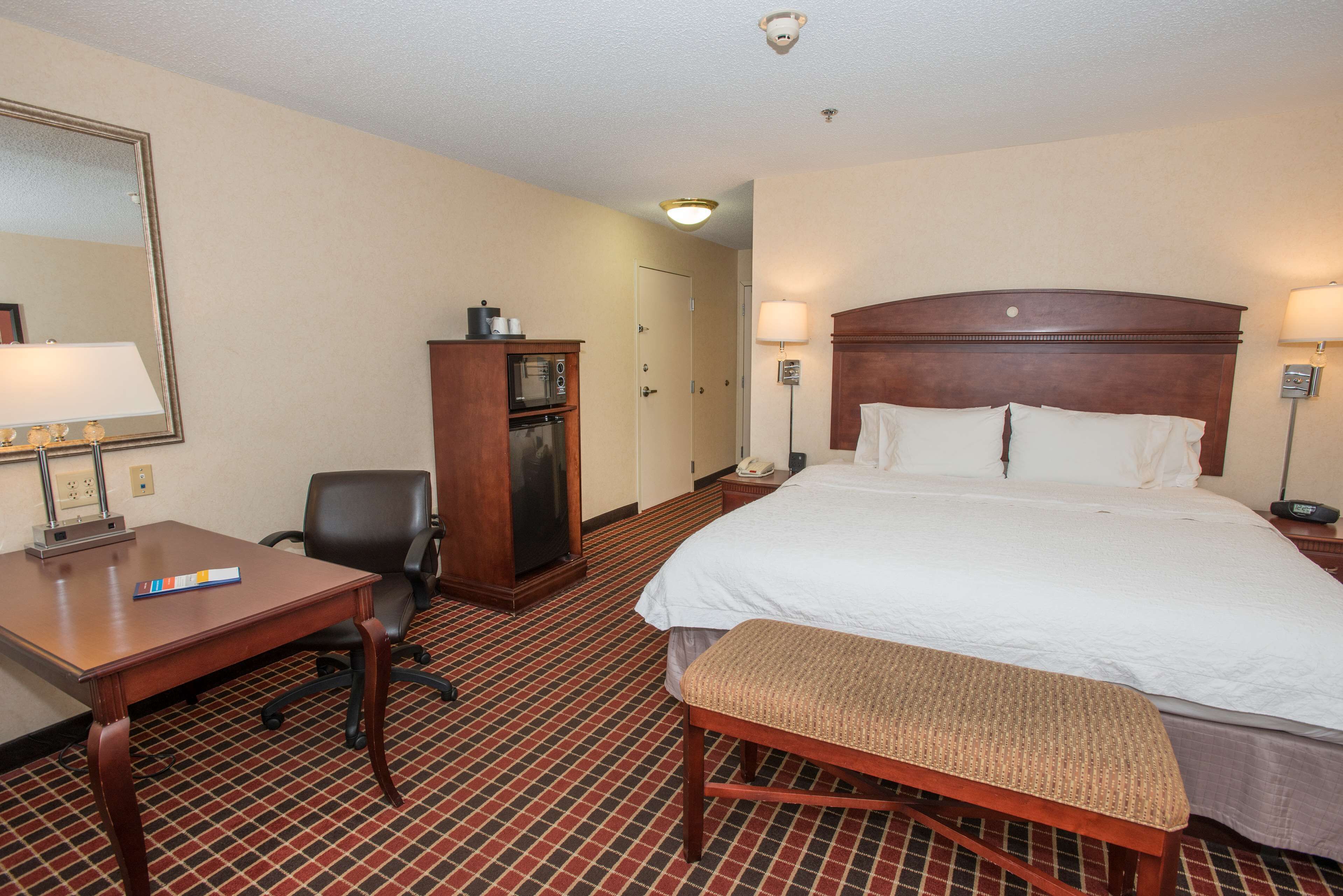A hotel room with a large bed, a desk, a chair, a small cabinet, and a bench.