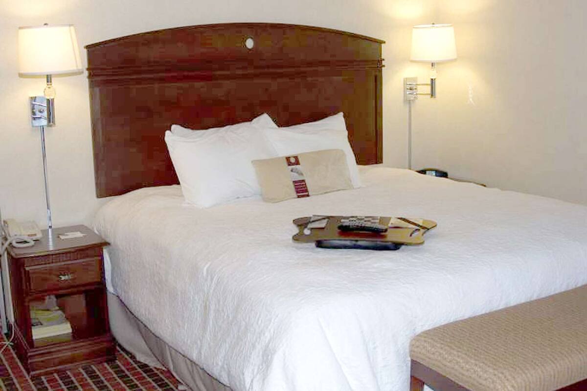 A neatly made bed with white linens, a wooden headboard, two bedside lamps, a bedside table with a phone, and a tray with remote controls.