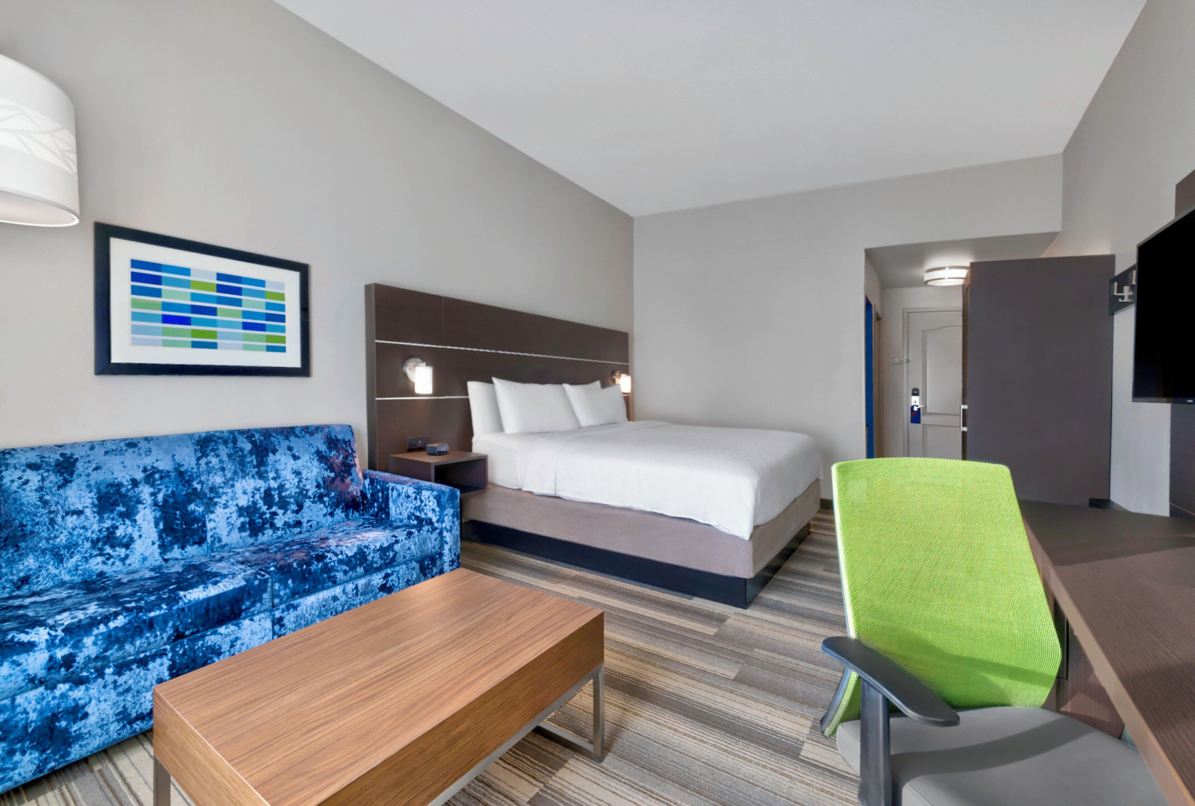 A hotel room with a bed, a blue sofa, a wooden table, a green chair, a framed picture, and a television.