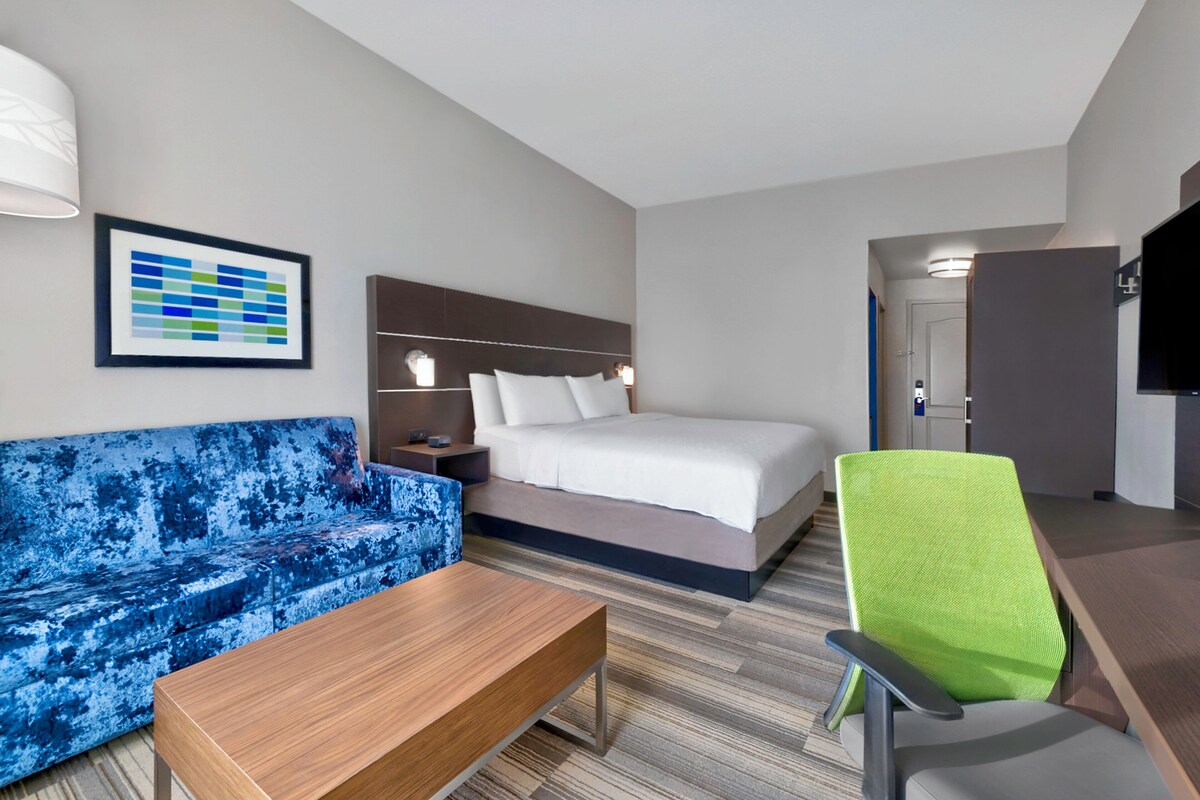 A hotel room with a bed, a blue sofa, a wooden table, a green chair, a framed picture, and a television.