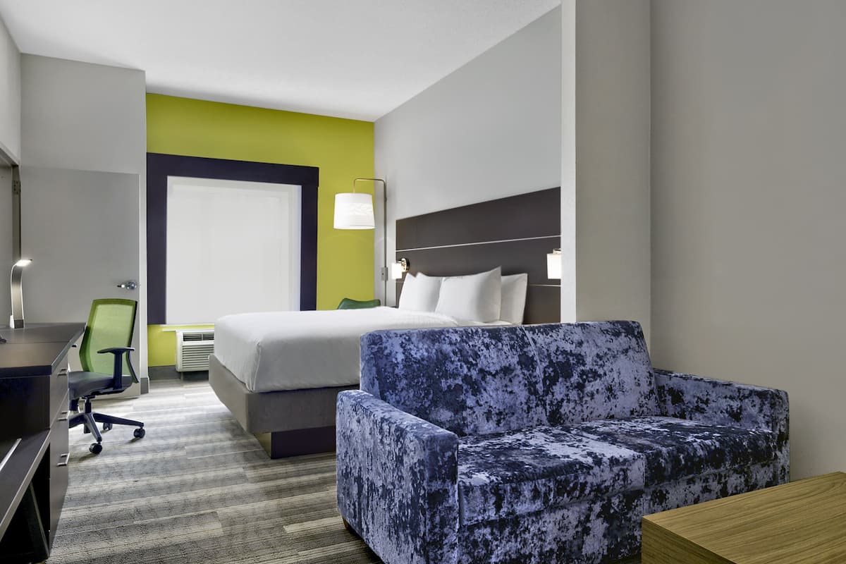 Une chambre d’hôtel avec un lit, un fauteuil bleu, un bureau avec une chaise et une table de chevet.