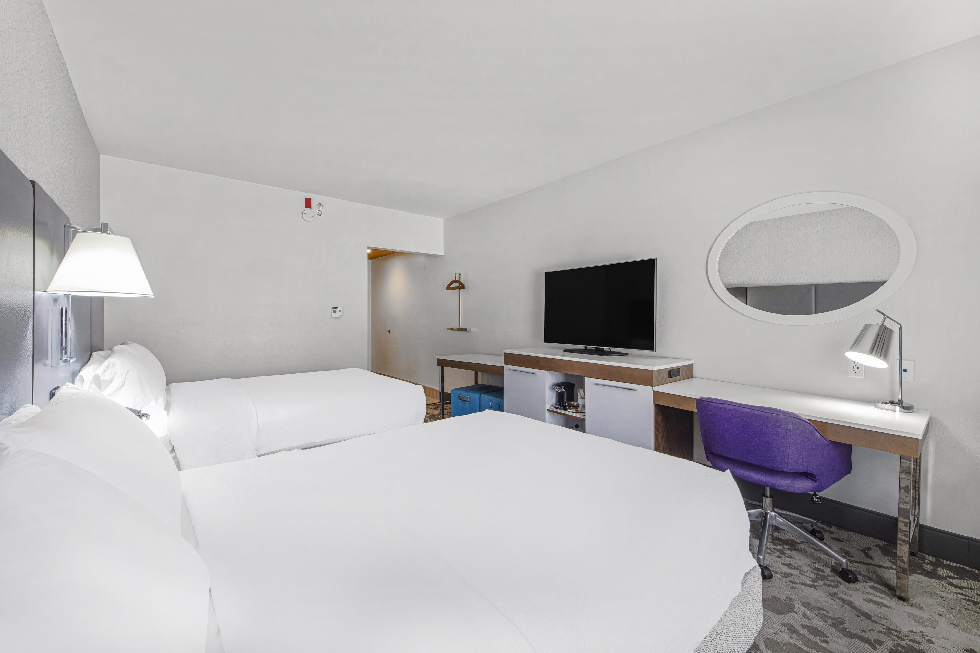 A hotel room with two beds, a desk with a computer, a TV, a mirror, and a lamp.