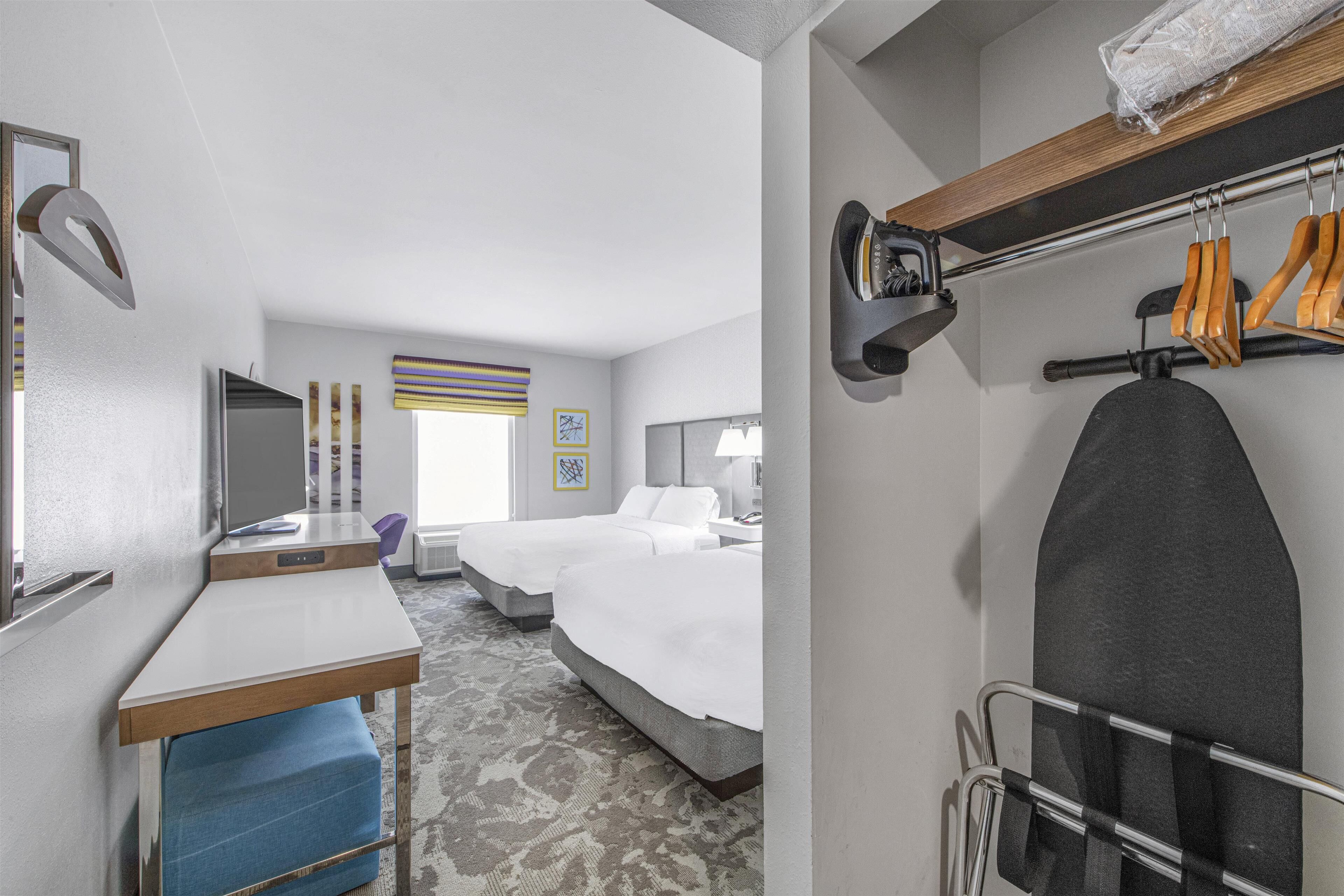 A hotel room with a desk, two beds, a chair, a TV, a mirror, and a coat rack.