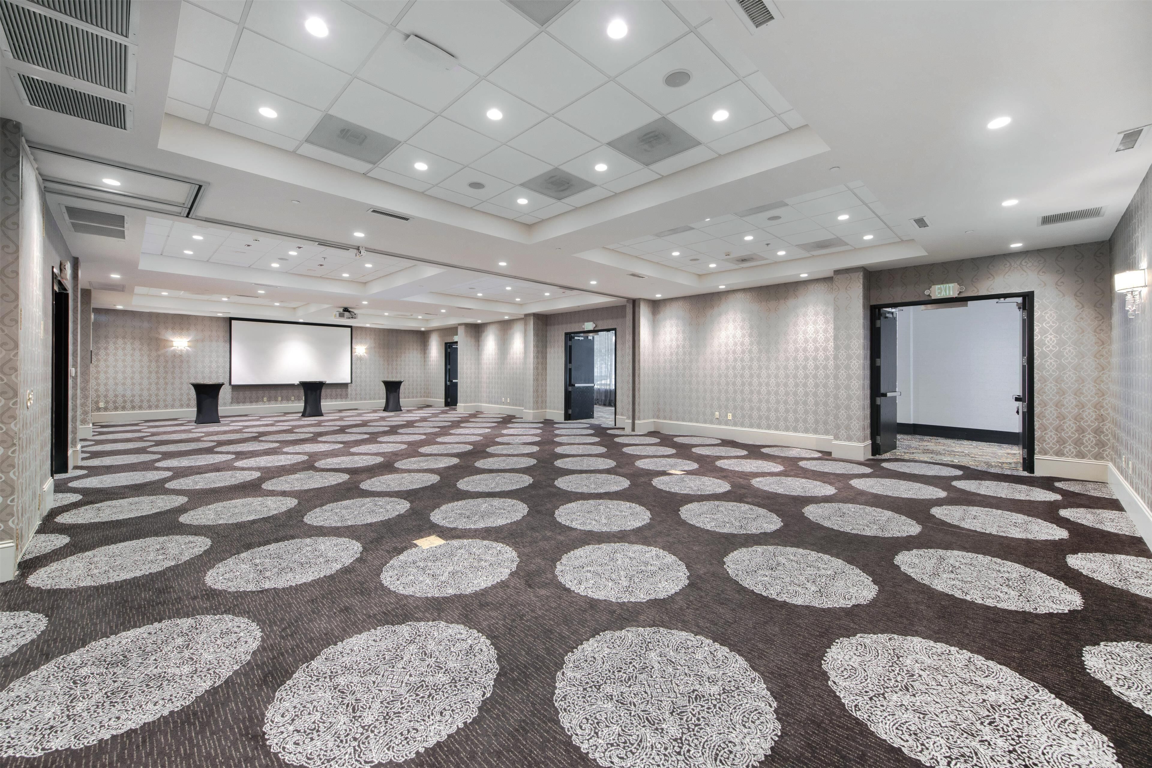 A spacious conference room with a patterned carpet, multiple doors, and a projector screen.