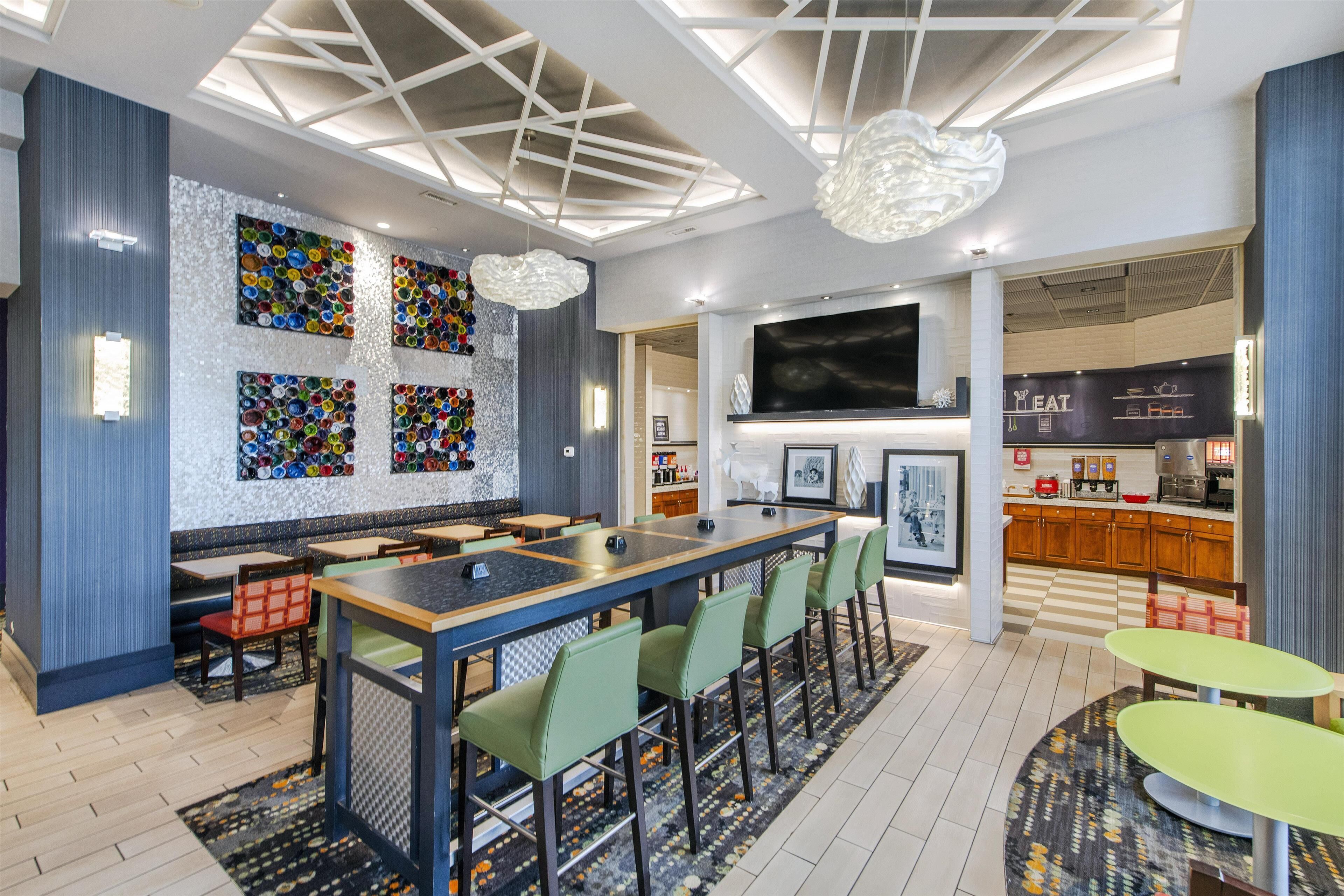 A modern bar area with a long counter, green chairs, and a colorful wall decoration.