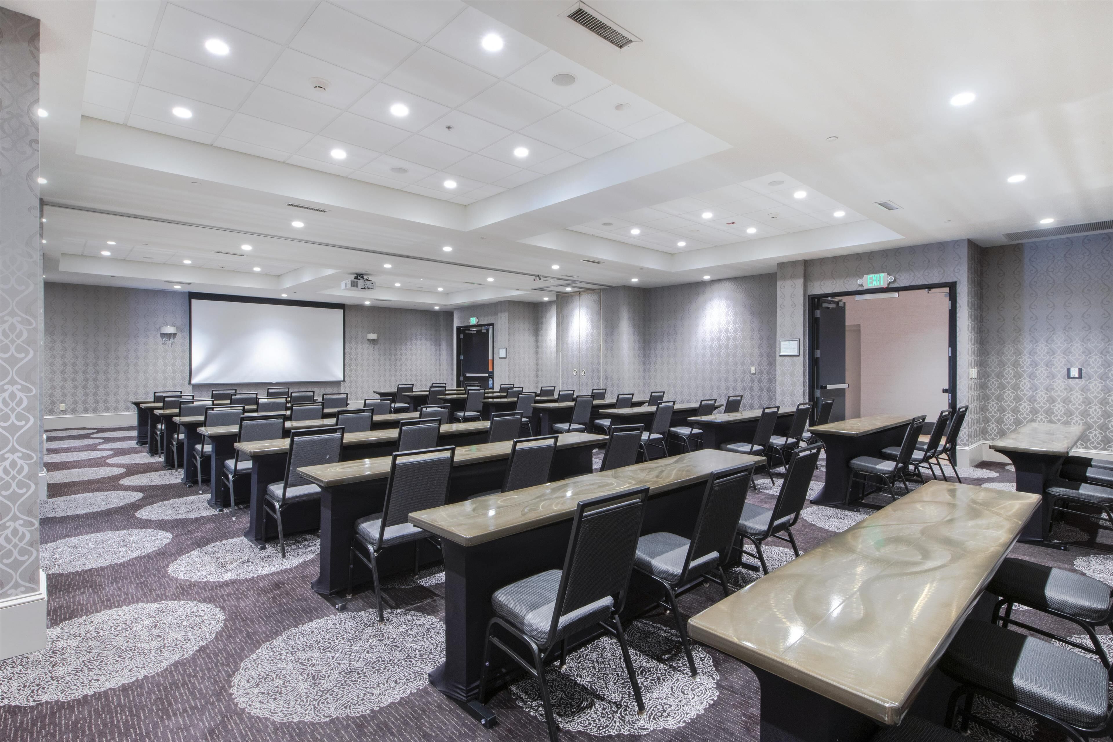 Un salón de conferencias con varias mesas y sillas, una pantalla de proyector y suelo alfombrado.
