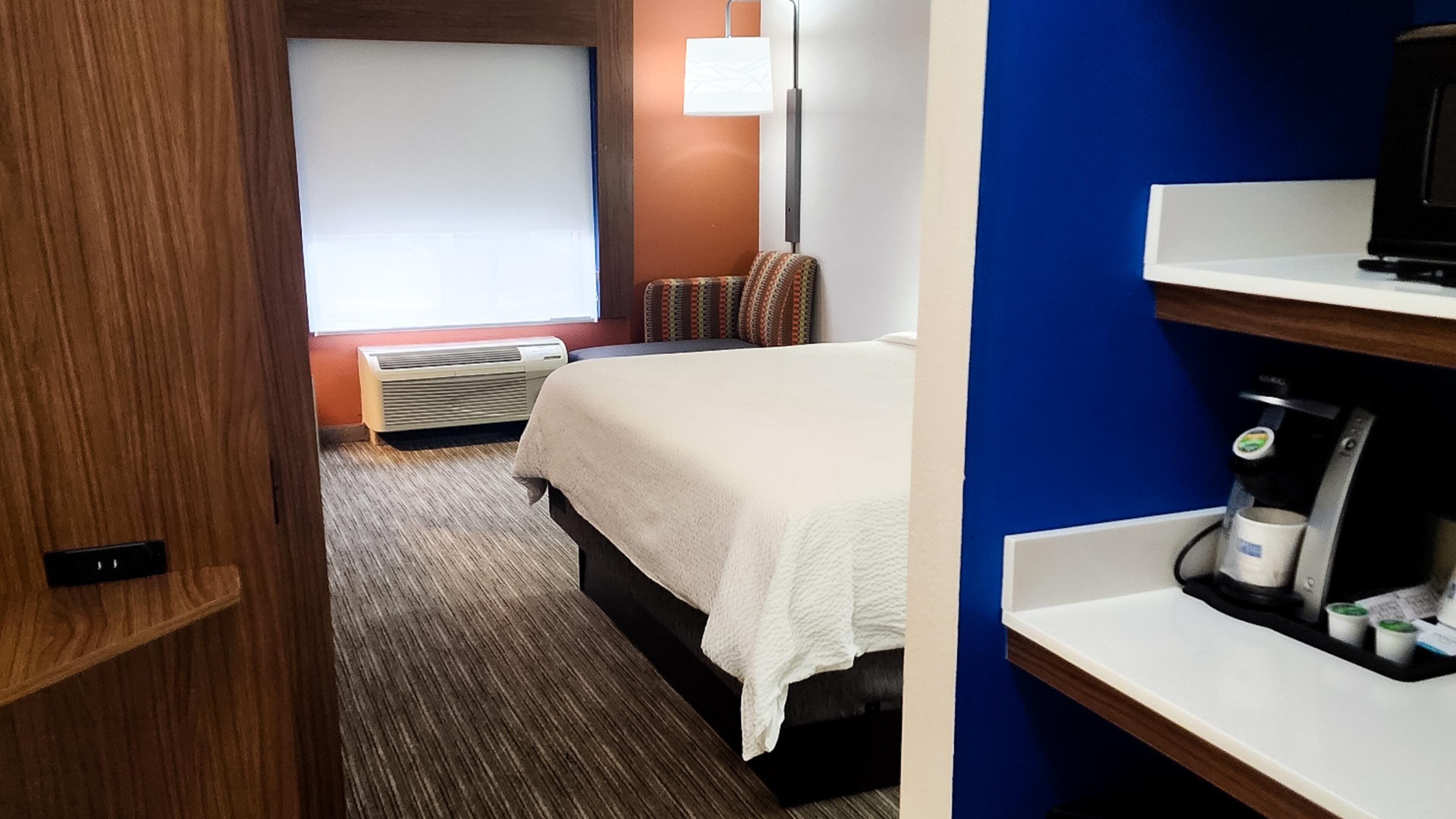 A hotel room with a bed, a desk with a coffee machine, a TV, a window with blinds, and a wall-mounted lamp.
