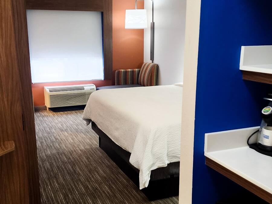 A hotel room with a bed, a desk with a coffee machine, a TV, a window with blinds, and a wall-mounted lamp.