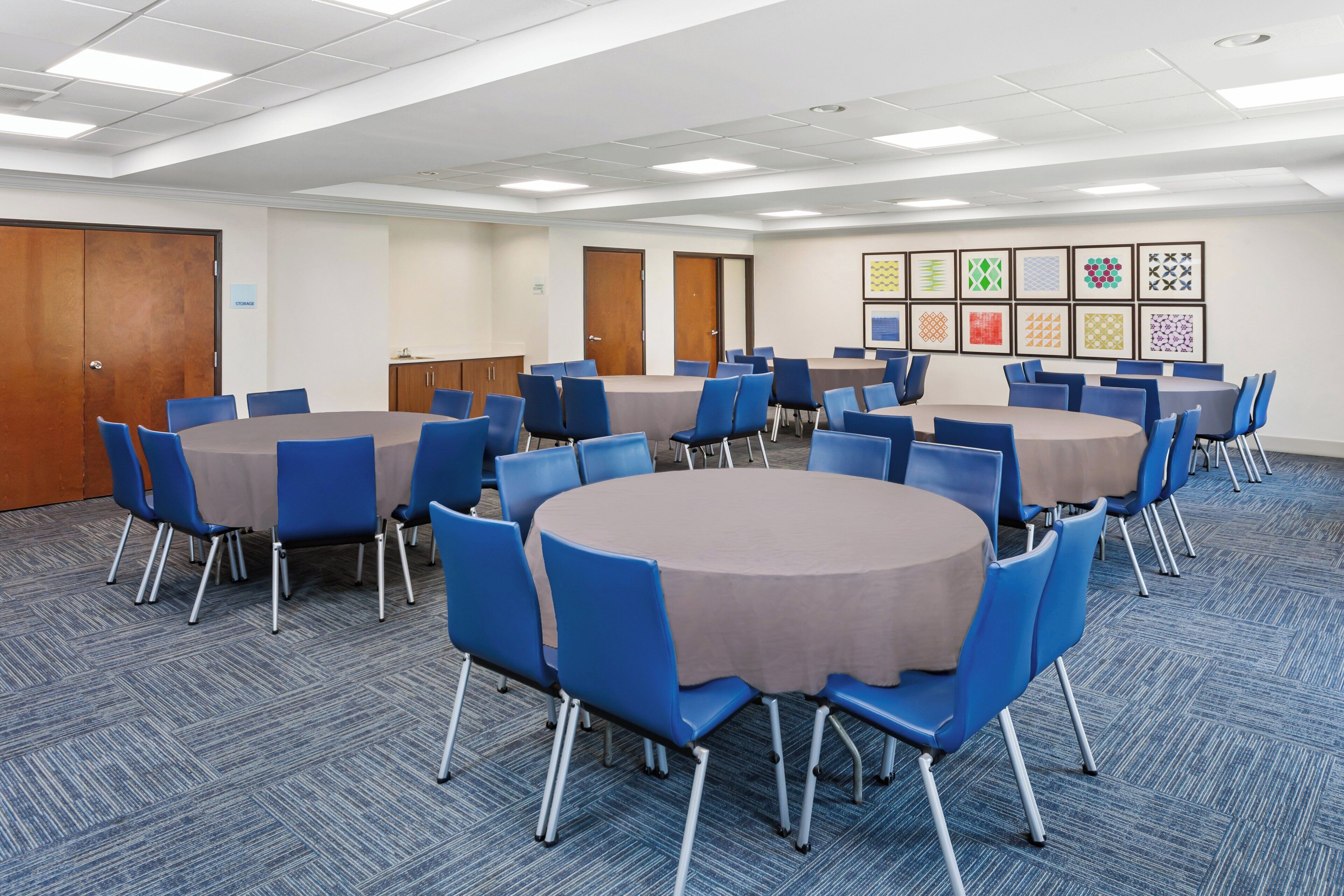 A conference room with round tables and blue chairs, carpeted floor, and framed artwork on the wall.