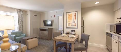 A modern hotel room with a dining area, a living room with a sofa and a TV, and a kitchen with a microwave and dishwasher.