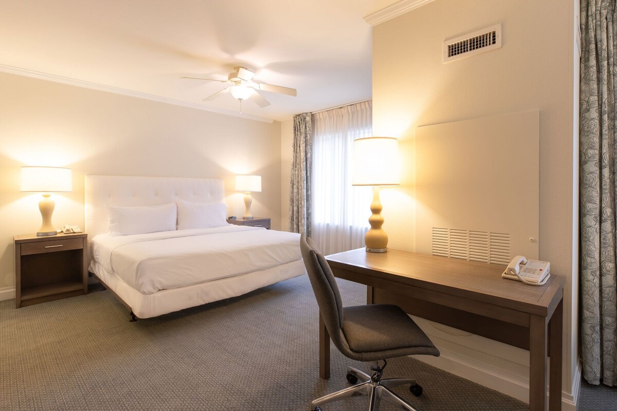 Ein Hotelzimmer mit Bett, Schreibtisch, Stuhl, zwei Nachttischlampen, Deckenventilator und Telefon.