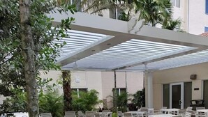 An outdoor patio area with white tables and chairs, surrounded by lush greenery and a pergola structure. - Homewood Suites by Hilton Palm Beach Gardens (Palm Beach Gardens)