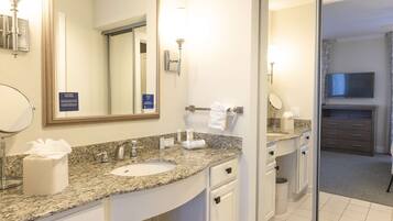 A hotel bathroom with a granite countertop sink, a large mirror, a trash can, and a television in the adjacent room.