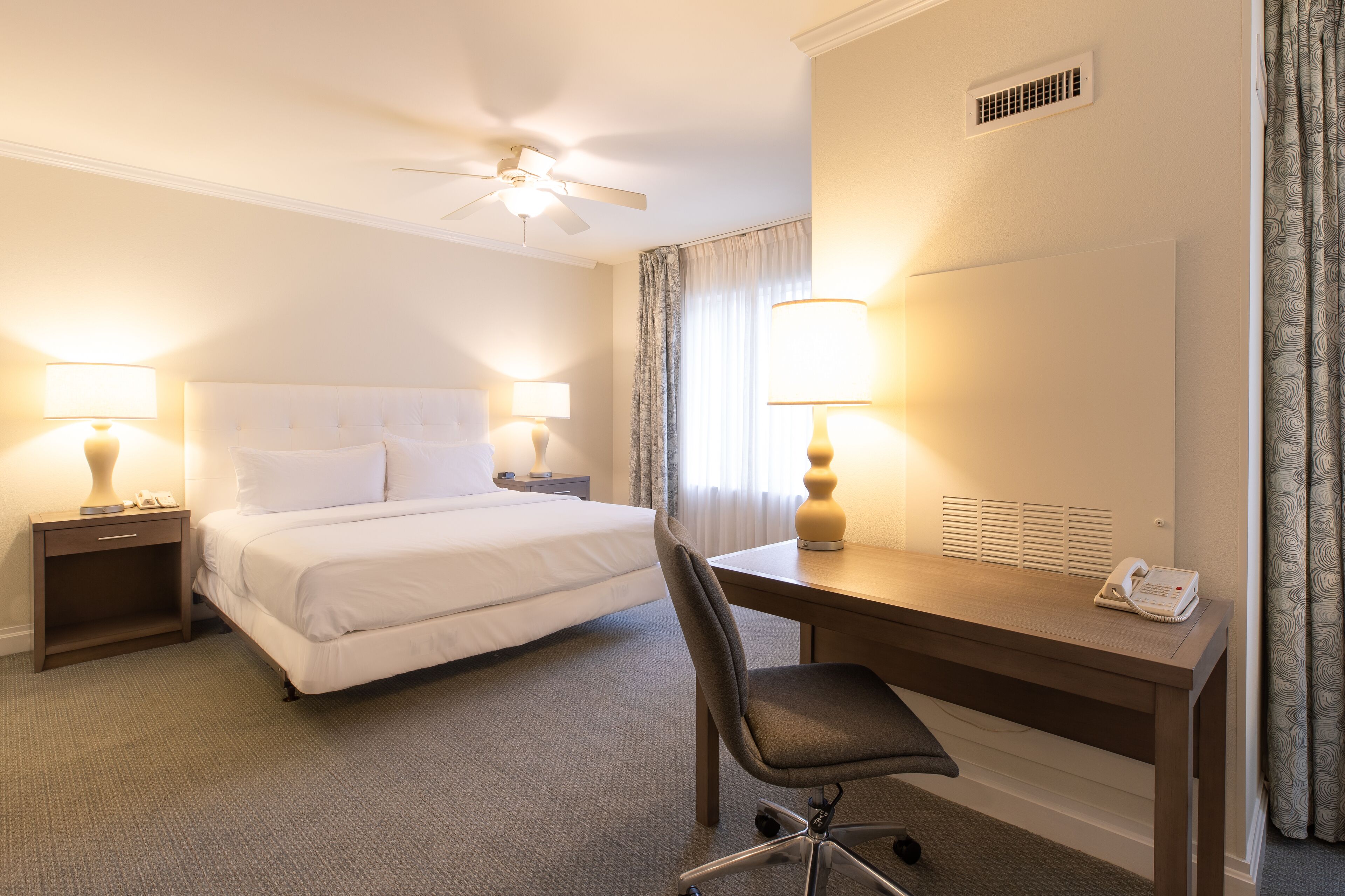 A hotel room with a bed, desk, chair, lamps, and a ceiling fan.