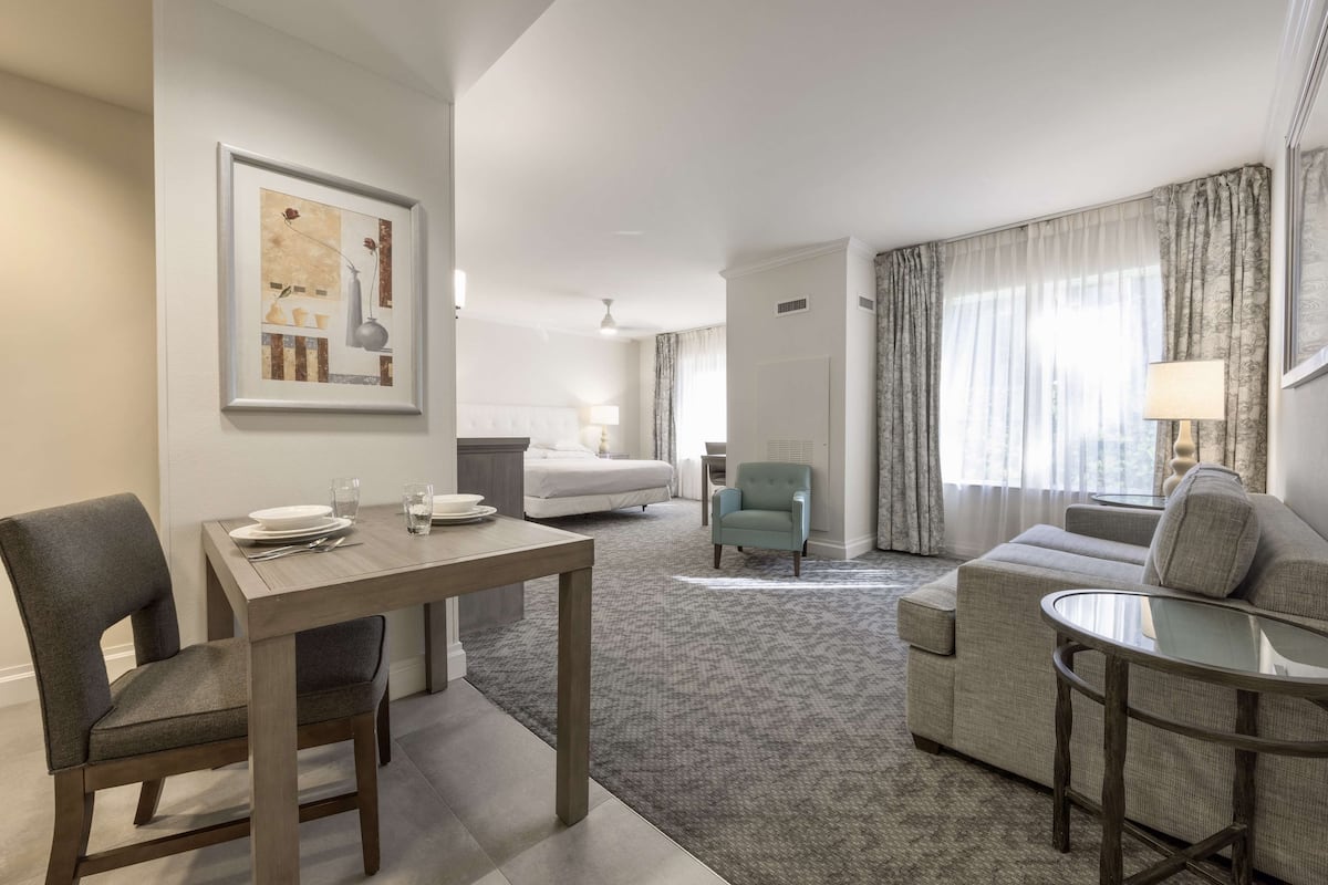 Une chambre d’hôtel comprenant une table à manger, un canapé, un petit fauteuil, un lit et une grande fenêtre avec des rideaux.