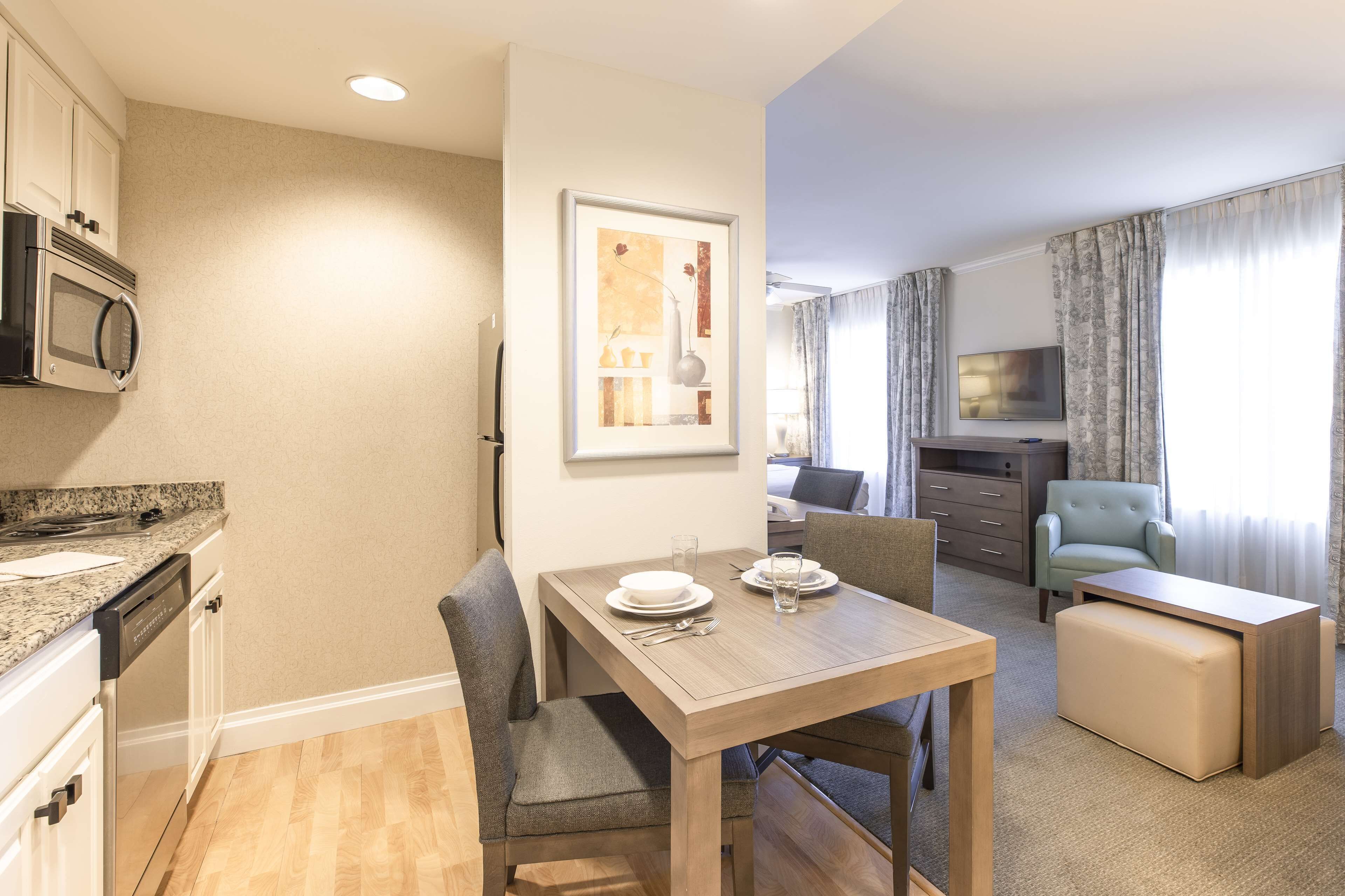 A modern kitchen with a built-in microwave, a dining area with a wooden table and grey upholstered chairs, and a living area with a sofa and a television.