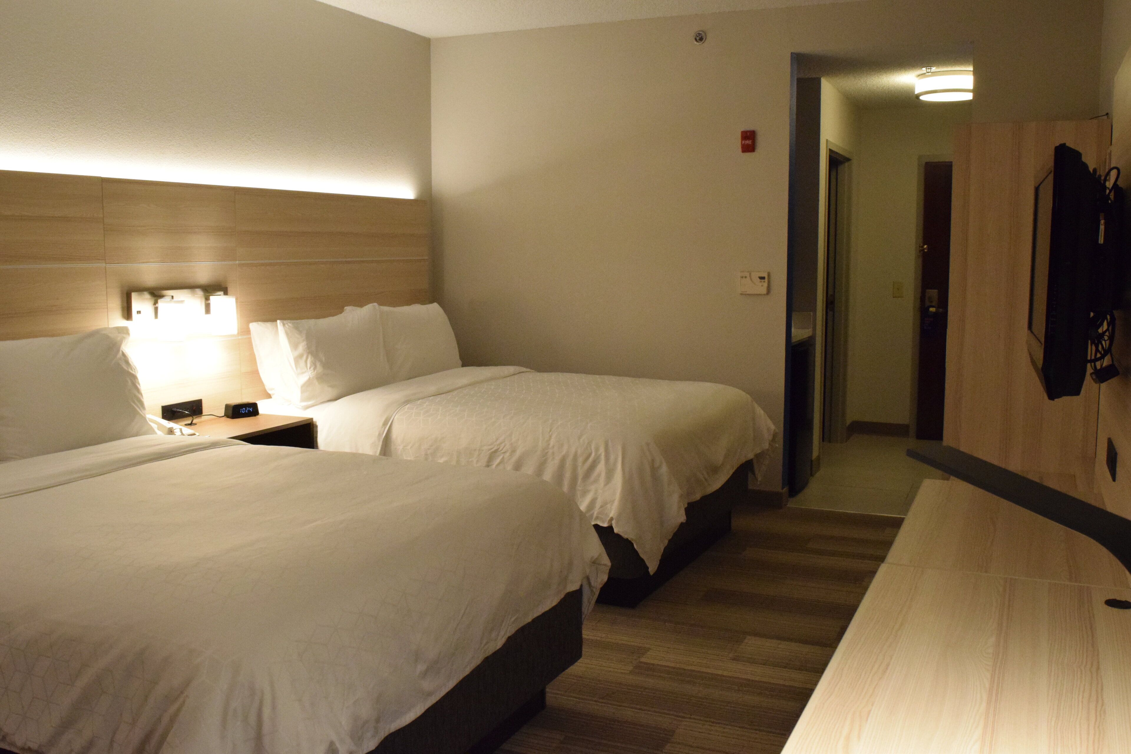 Een hotelkamer met twee bedden, een nachtkastje met een lamp, een houten hoofdbord, een aan de muur bevestigde televisie en een deur naar een gang.