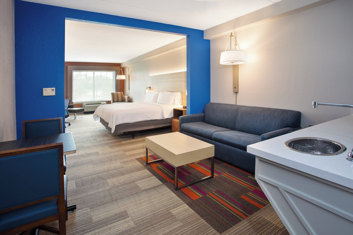 A hotel room with a bed, a sofa, a desk, a chair, a small table, a sink, and a window with a view.