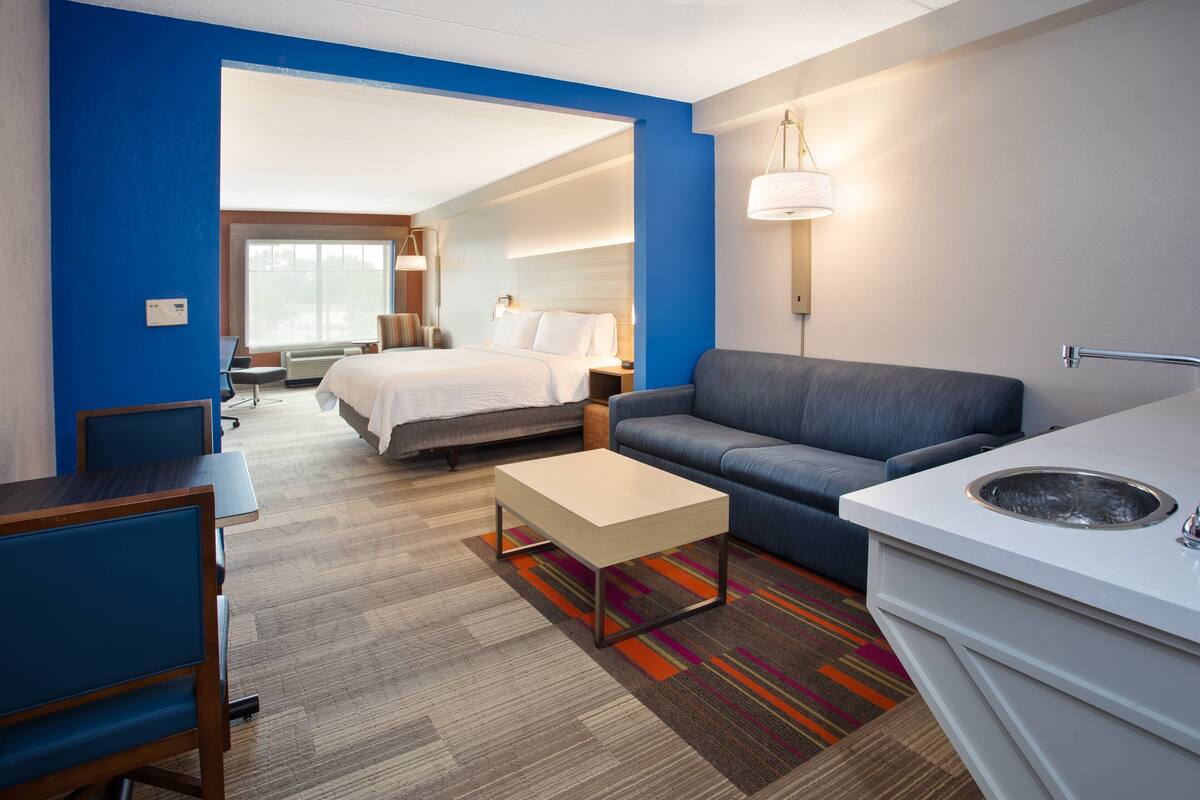 A hotel room with a bed, a sofa, a desk, a chair, a small table, a sink, and a window with a view.